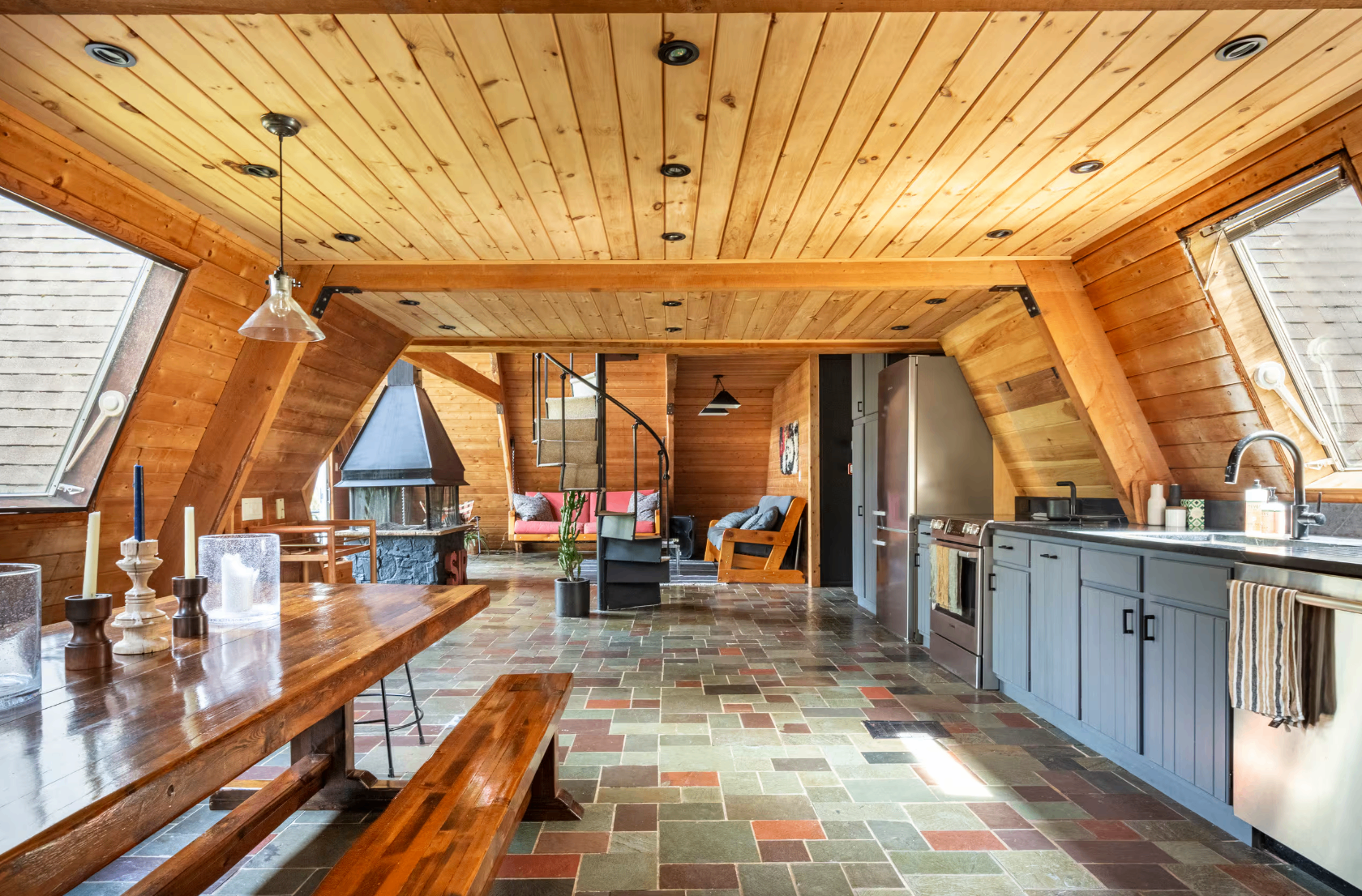 Open-plan A-frame interior with wood-paneled ceiling, kitchen, dining table, and spiral staircase