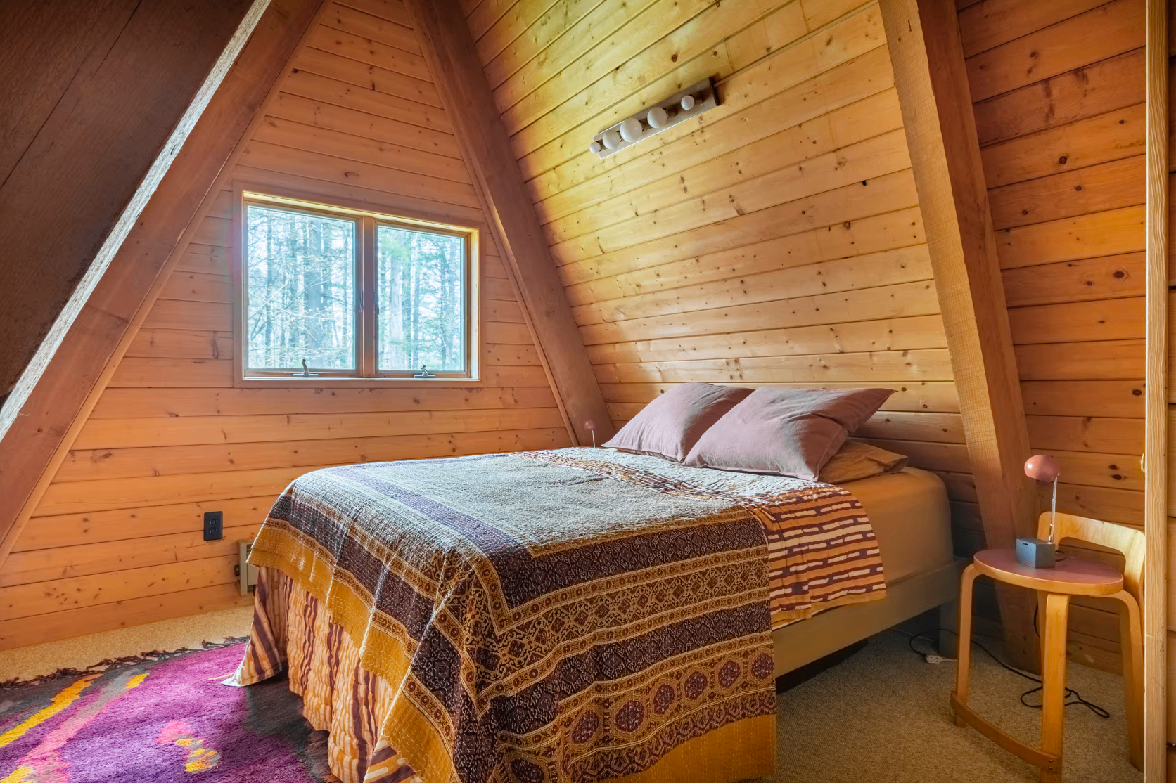 A-frame bedroom with warm wood walls, patterned quilt, and forest view window
